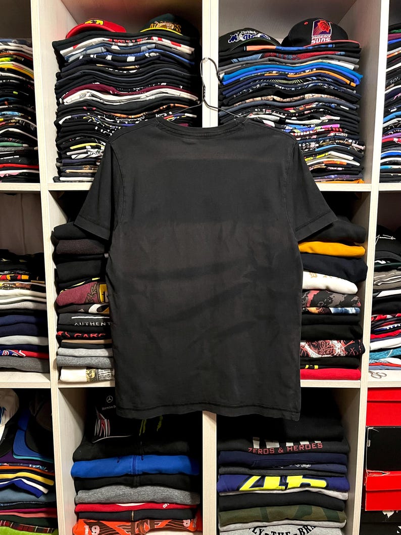 May include: A black t-shirt hangs in front of a shelf filled with folded clothing and hats. The t-shirt is plain and appears to be made of a soft material. The shelf is divided into compartments, each holding stacks of various items.