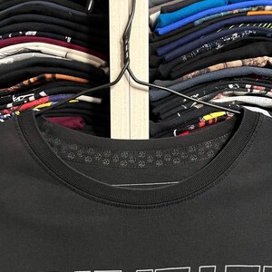 May include: A black t-shirt with the word "AMMS+E" printed in white. The shirt is hanging on a black hanger, with a closet full of folded shirts in the background.