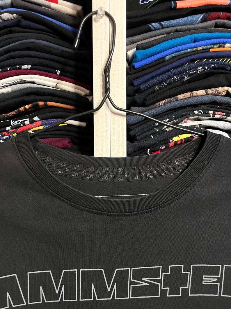 May include: A black t-shirt with the word "AMMS+E" printed in white. The shirt is hanging on a black hanger, with a closet full of folded shirts in the background.