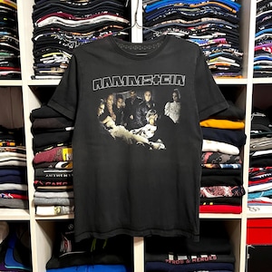 May include: Black Rammstein band t-shirt with a graphic of the band members. The band's name is printed in white block letters across the top. The shirt is hanging on a metal hanger in front of a shelf of folded clothing.