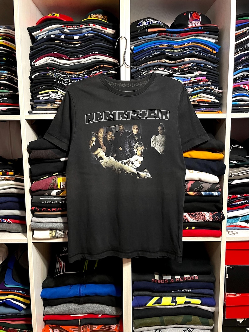 May include: Black Rammstein band t-shirt with a graphic of the band members. The band's name is printed in white block letters across the top. The shirt is hanging on a metal hanger in front of a shelf of folded clothing.