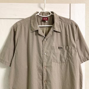 May include: Tan short-sleeve button-down shirt with a small, repeating black dot pattern. The shirt has a chest pocket and a collar. The Oakley logo is visible on the collar and pocket.