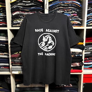 May include: Black t-shirt with the text "RAGE AGAINST THE MACHINE" in white. The shirt features a white graphic of two figures in a circle. The shirt is hanging on a hanger in front of a shelf filled with folded clothing and hats.