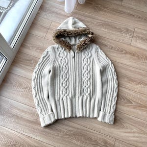 May include: A light grey cable-knit hooded cardigan with a faux fur-lined hood. The cardigan features a full zip and ribbed cuffs and hem. The cable-knit pattern is visible on the sleeves and front panels. It is on a wooden floor.