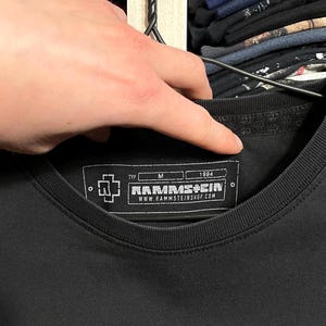 May include: Black crew neck t-shirt with the band name "RAMMSTEIN" printed in white. The shirt features a tag with the band's logo, size "M", and the year "1984". The website address "WWW.RAMMSTEINSHOP.COM" is also visible on the tag.