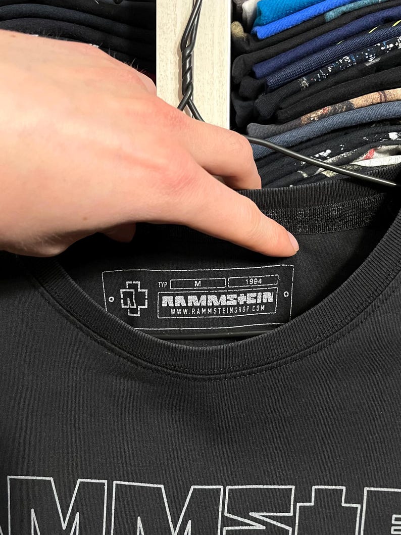 May include: Black crew neck t-shirt with the band name "RAMMSTEIN" printed in white. The shirt features a tag with the band's logo, size "M", and the year "1984". The website address "WWW.RAMMSTEINSHOP.COM" is also visible on the tag.