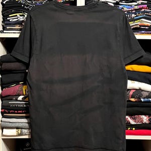May include: A black short-sleeved t-shirt, displayed on a hanger. The shirt is a solid color and appears to be made of a soft material. The background shows shelves filled with folded clothing.