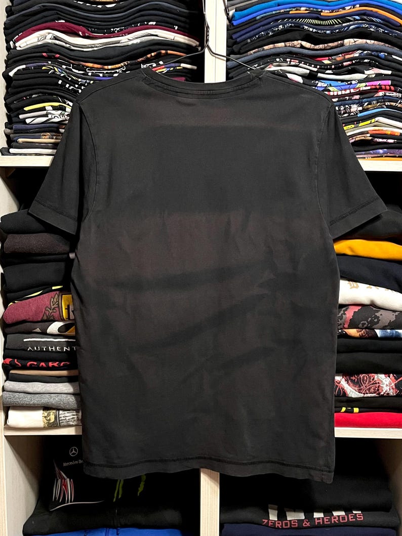 May include: A black short-sleeved t-shirt, displayed on a hanger. The shirt is a solid color and appears to be made of a soft material. The background shows shelves filled with folded clothing.