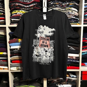 May include: Black t-shirt featuring a graphic of a crowd holding a banner that reads "SYSTEM OF A DOWN" in red letters. The shirt is hanging on a rack in front of a shelf filled with folded clothing.