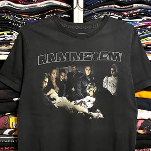 May include: Black t-shirt featuring the band name "RAMMSTEIN" in white block letters above a grayscale image of the band members. The image shows the band members in a group, with a central figure lying down.
