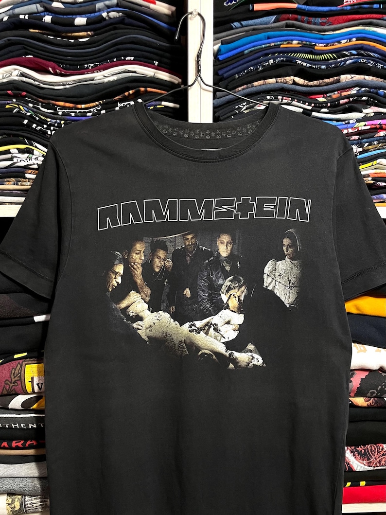 May include: Black t-shirt featuring the band name "RAMMSTEIN" in white block letters above a grayscale image of the band members. The image shows the band members in a group, with a central figure lying down.