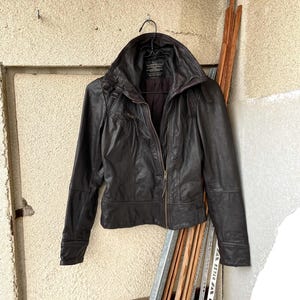 Peut inclure: Veste en cuir marron foncé avec un col montant et une fermeture éclair. La veste est suspendue à un cintre noir contre un mur beige texturé. La veste a une coupe ajustée et des manches longues.