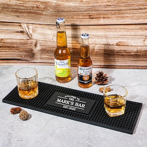 Puede incluir: Una alfombrilla de bar negra con dos vasos de líquido ámbar, dos botellas de cerveza y un letrero que dice "WELCOME TO THE MARK'S BAR EST. 2025". Las botellas están etiquetadas como "Clavelito" y "Coronita Extra".