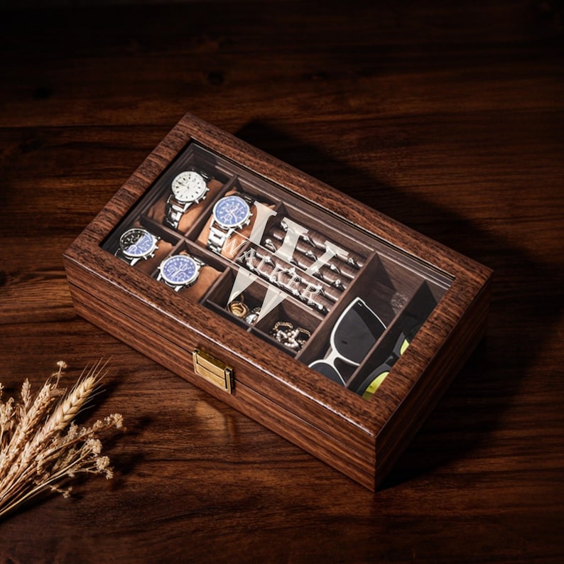 Op de afbeelding: Een houten horloge- en accessoire-organizer met een doorzichtig glazen bovenkant. De binnenkant heeft compartimenten voor horloges, ringen en zonnebrillen. De organizer is gemaakt van donker hout en heeft een goudkleurige sluiting.