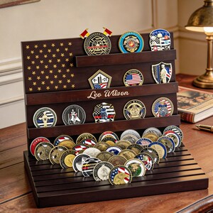 May include: A dark brown wooden coin display case with multiple shelves, holding a collection of military and commemorative coins. The case features an American flag design and the name "Leo Wilson". The coins are in various colors and designs.