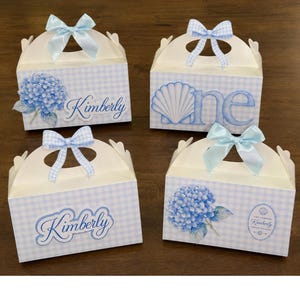 May include: Four white cardboard favour boxes with light blue gingham patterns, each topped with a blue ribbon bow. The boxes feature printed designs of blue hydrangea flowers, a seashell, and the name Kimberly or the word one in blue script.