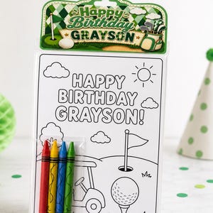 May include: A golf-themed birthday coloring set with the text "Happy Birthday Grayson!" includes a coloring page, crayons, and a decorative topper. The coloring page features a golf course scene with a golf cart, ball, and flag.