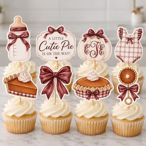 May include: A set of eight decorative cupcake toppers featuring illustrations of a baby bottle, a sign reading A LITTLE CUTIE PIE IS ON THE WAY, a pumpkin with a monogram, a baby romper, a slice of pie, a large bow, a whole pie, and a rattle.