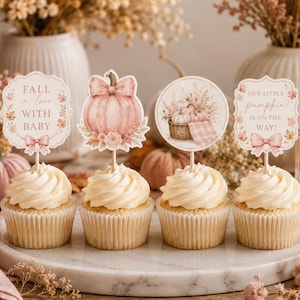 Könnte beinhalten: Vier Cupcakes mit dekorativen Papieraufsteckern, die rosa Kürbisse, Blumenmotive und Schleifen zeigen. Der Text lautet FALL in love WITH BABY und OUR LITTLE pumpkin IS ON THE WAY! Die Aufstecker stecken auf weißen Stäbchen im Frosting.