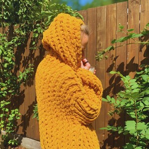 May include: A mustard yellow hooded sweater made of thick, chunky knit yarn. The sweater has a large hood and long sleeves. The person wearing the sweater is standing outside near a wooden fence and green foliage.