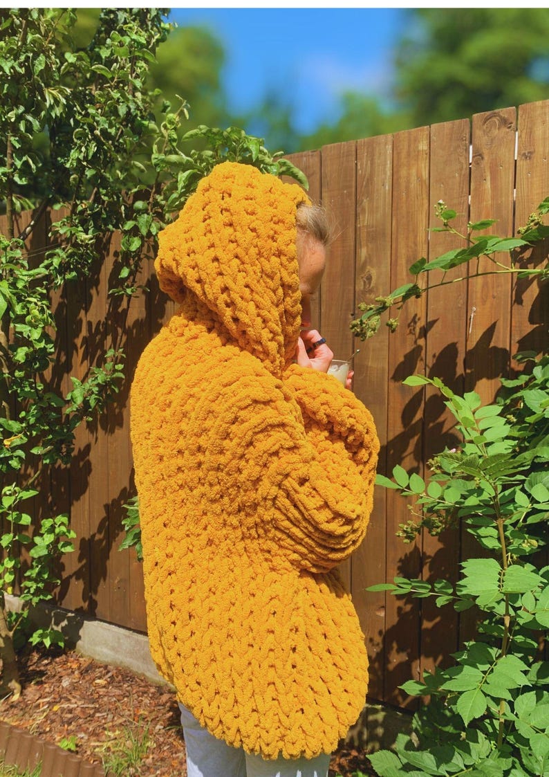 May include: A mustard yellow hooded sweater made of thick, chunky knit yarn. The sweater has a large hood and long sleeves. The person wearing the sweater is standing outside near a wooden fence and green foliage.