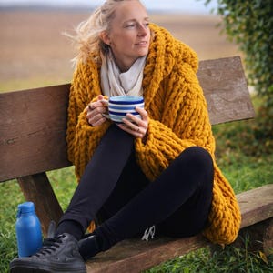 May include: A woman wearing a mustard yellow hooded knit sweater, black leggings, and black sneakers sits on a wooden bench. She holds a blue and white striped mug. A blue water bottle sits nearby. The background is a field and trees.