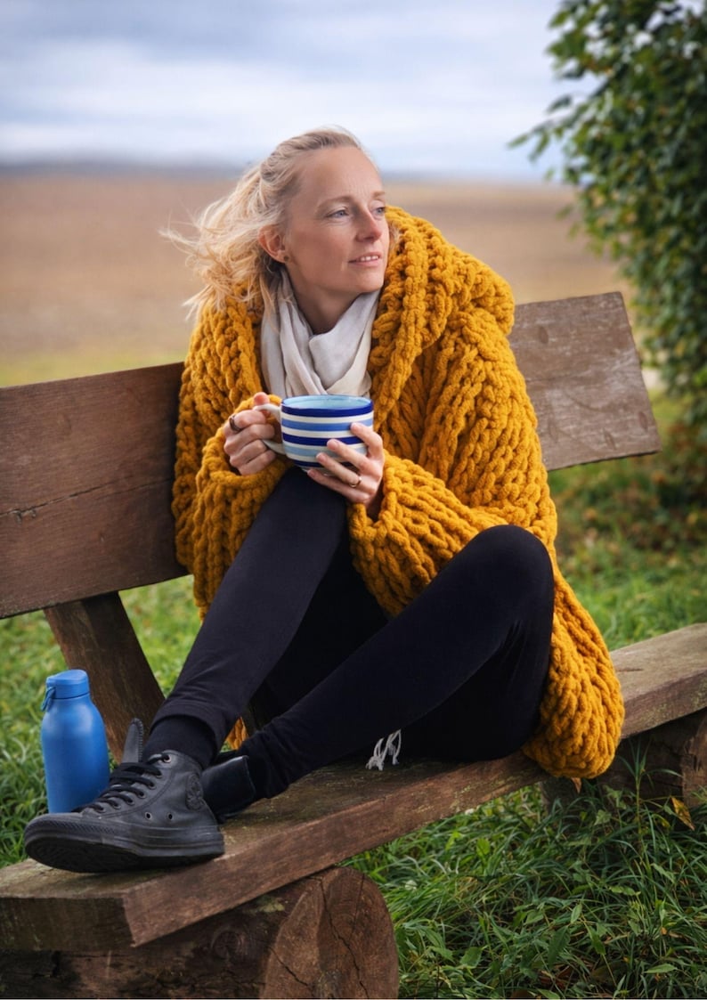 May include: A woman wearing a mustard yellow hooded knit sweater, black leggings, and black sneakers sits on a wooden bench. She holds a blue and white striped mug. A blue water bottle sits nearby. The background is a field and trees.
