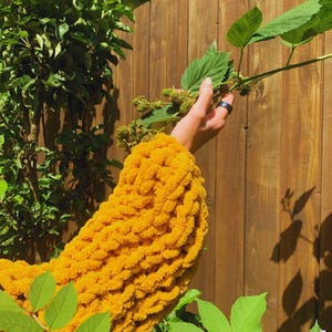 May include: A close-up of a bright yellow, chunky knit sweater sleeve. The sleeve is made of thick yarn and has a textured appearance. The background includes a wooden fence, green leaves, and a hand holding a plant with green berries.