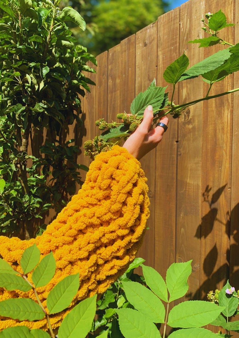 May include: A close-up of a bright yellow, chunky knit sweater sleeve. The sleeve is made of thick yarn and has a textured appearance. The background includes a wooden fence, green leaves, and a hand holding a plant with green berries.