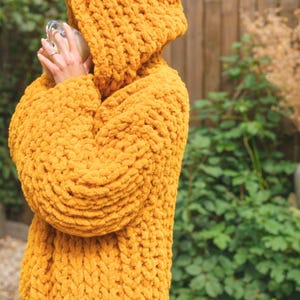 May include: Mustard yellow hooded sweater. The chunky knitwear features a large hood and long sleeves. The sweater is made of a soft, textured material. The image is taken outdoors with a blurred background of greenery and a wooden fence.