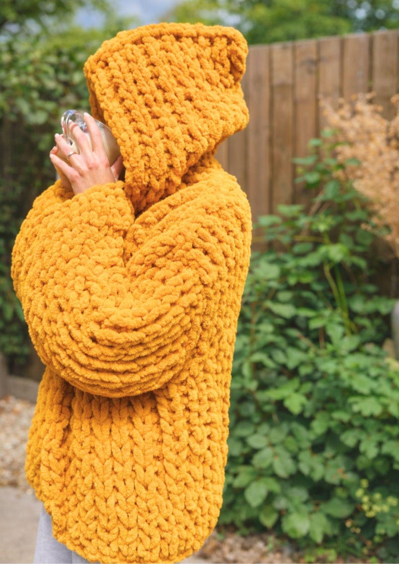 May include: Mustard yellow hooded sweater. The chunky knitwear features a large hood and long sleeves. The sweater is made of a soft, textured material. The image is taken outdoors with a blurred background of greenery and a wooden fence.