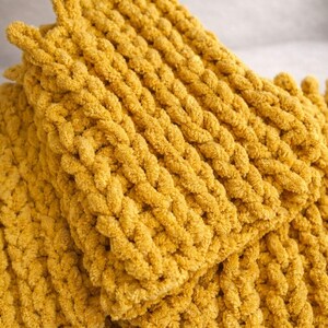May include: Close-up of a stack of two mustard yellow knitted blankets. The blankets are made of a thick, textured yarn, with a braided pattern. The edges of the blankets have a decorative, looped trim. The blankets are soft and cozy.