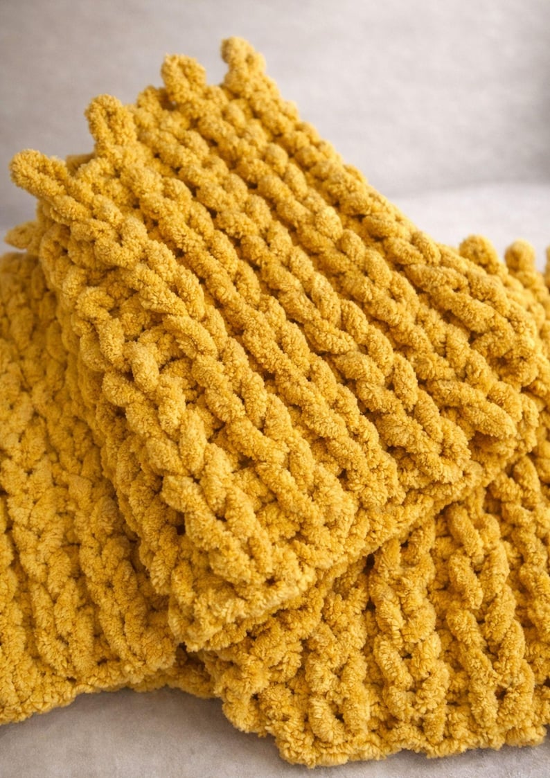 May include: Close-up of a stack of two mustard yellow knitted blankets. The blankets are made of a thick, textured yarn, with a braided pattern. The edges of the blankets have a decorative, looped trim. The blankets are soft and cozy.