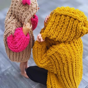 May include: Two children's hooded sweaters. One is tan with pink accents, the other is mustard yellow. Both are made of a chunky knit material. The children are wearing the sweaters.