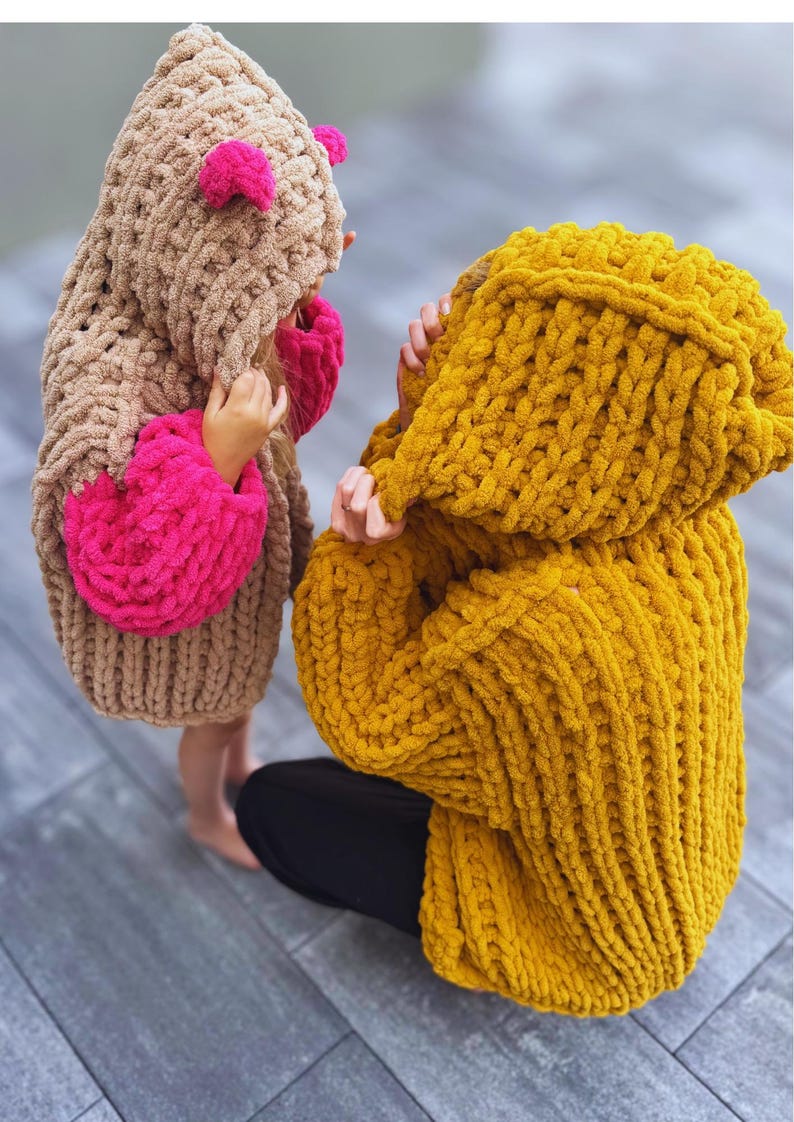 May include: Two children's hooded sweaters. One is tan with pink accents, the other is mustard yellow. Both are made of a chunky knit material. The children are wearing the sweaters.
