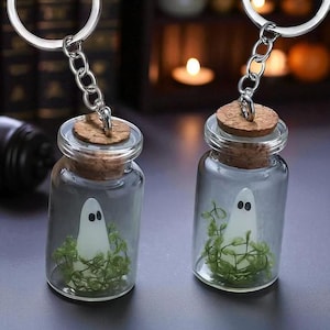Peut inclure: Deux porte-clés en forme de bouteille en verre avec un bouchon en liège. Chaque bouteille contient un petit fantôme blanc et du feuillage vert. Les porte-clés sont attachés à des anneaux en métal argenté. L'arrière-plan est flou.