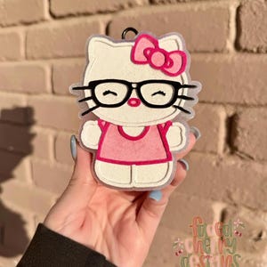 May include: A white cat-shaped charm with a pink bow, glasses, and a pink outfit. The charm has a black outline and a small loop at the top. The charm is held in a hand.