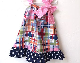 May include: A colorful patchwork dress with a pink polka dot bow and straps. The dress features a navy blue polka dot ruffle at the hem. The dress is hanging on a white hanger.