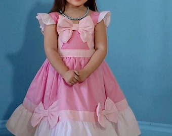 Puede incluir: Un vestido rosa con detalles blancos y lazos grandes. El vestido presenta un lazo en la cintura y en la falda. El conjunto se completa con una diadema blanca y un collar de perlas. El fondo es azul liso.