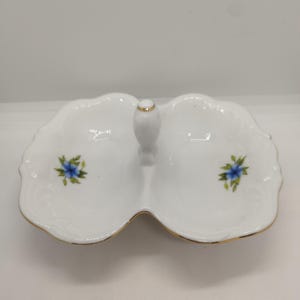 Peut inclure: Plat de service en porcelaine blanche divisé en deux compartiments avec une poignée. Le plat a un bord festonné, des motifs floraux bleus et une bordure dorée. La poignée est également ornée d'or.