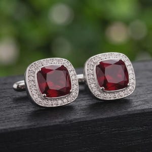 Puede incluir: Un par de gemelos cuadrados con una piedra central de color rojo intenso en talla cojín, rodeada por un halo de pequeñas piedras claras en talla redonda, montados en un marco de metal plateado con cierre de palanca estándar.