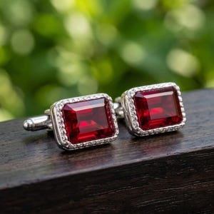 May include: A pair of rectangular cufflinks featuring deep red, emerald-cut stones set in silver-toned metal frames. Each stone is surrounded by a border of small, clear, reflective accents, with a polished metallic toggle mechanism on the back.