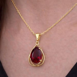 May include: A gold necklace with a teardrop-shaped, deep red gemstone pendant. The pendant is set in a delicate gold frame and hangs from a fine gold chain. The necklace is shown against a neutral background.