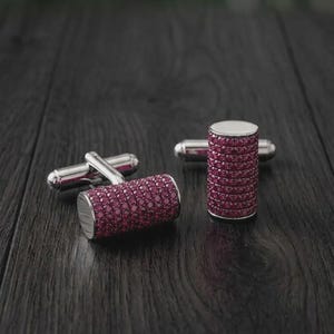 May include: A pair of cylindrical cufflinks featuring a polished silver-tone metal frame and a surface densely encrusted with small, vibrant red stones. One cufflink is positioned horizontally, while the other stands vertically on its flat end.