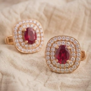 May include: A pair of gold-toned cufflinks featuring a central oval-cut deep red stone surrounded by two concentric rectangular halos of small, clear, sparkling white stones set in a metallic frame.