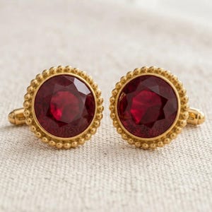 Puede incluir: Un par de gemelos redondos con piedras de color rojo intenso facetadas engastadas en metal dorado. Cada engaste circular está enmarcado por un borde de cuentas decorativo, con un mecanismo de palanca estándar visible en el lateral de la base.