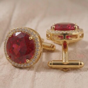 May include: A pair of gold-tone cufflinks featuring a large, round, faceted red stone at the center. The red stone is surrounded by a halo of small, clear, sparkling stones set in a gold-colored metal frame with a classic T-bar swivel closure.