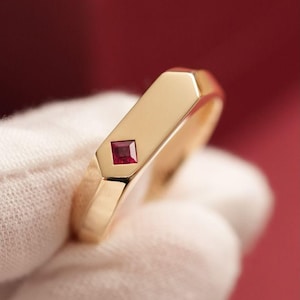 May include: A gold ring with a rectangular band and a square, red gemstone. The ring has a polished finish and a modern design. The gemstone is set in a bezel setting.