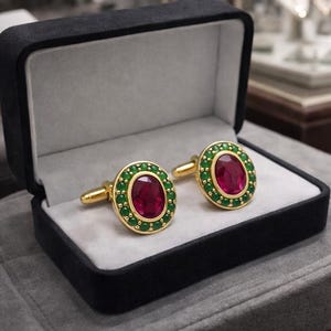 May include: A pair of gold cufflinks, each featuring a central oval ruby encircled by small green gems. They are displayed in a black velvet box with a grey lining, indicative of a high-end product.