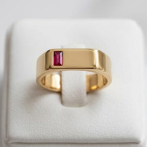 Könnte beinhalten: Ein polierter Goldring mit einem einzelnen, rechteckigen Rubin im Baguetteschliff, der bündig in die Metalloberfläche eingelassen ist. Der Ring hat ein glattes, breites Profil mit glänzender Oberfläche, die die tiefrote Farbe des Steins betont.